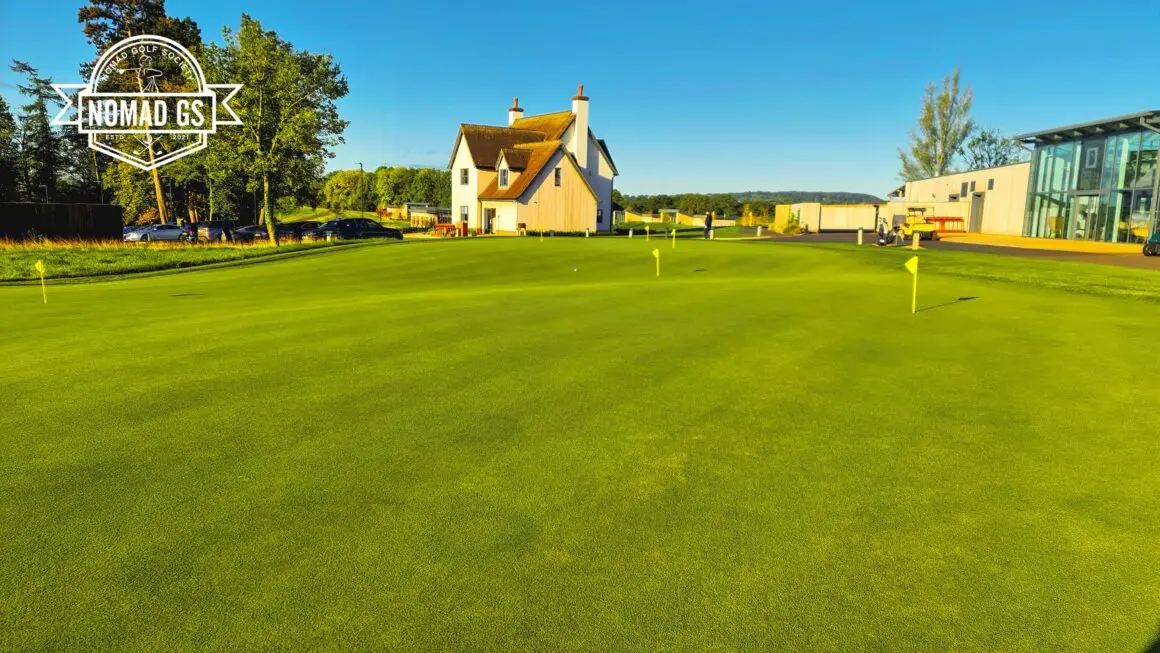 Large, immaculate putting green with yellow flags in front of JCB clubhouse buildings under a clear blue sky.