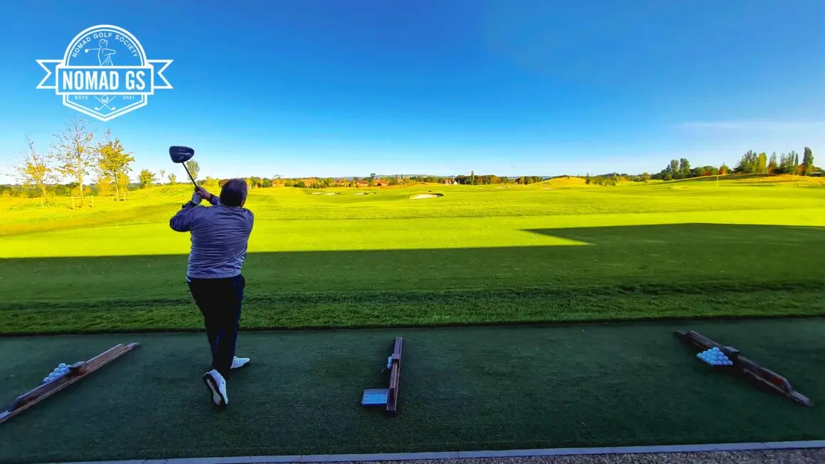 Golfer finishing a driver swing on JCB's grass driving range under clear blue sky, ball trays lined up beside him.